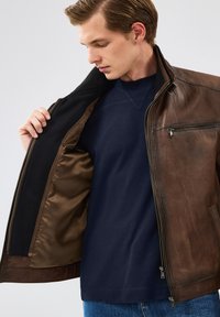 Veste en cuir marron avec une fermeture éclair à l'avant, un col côtelé et une doublure en satin beige. Un pull bleu marine est porté en dessous.