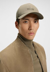 Khaki baseball cap with embroidered "BOSS" logo, worn by a model in a brown suede bomber jacket over a beige turtleneck.