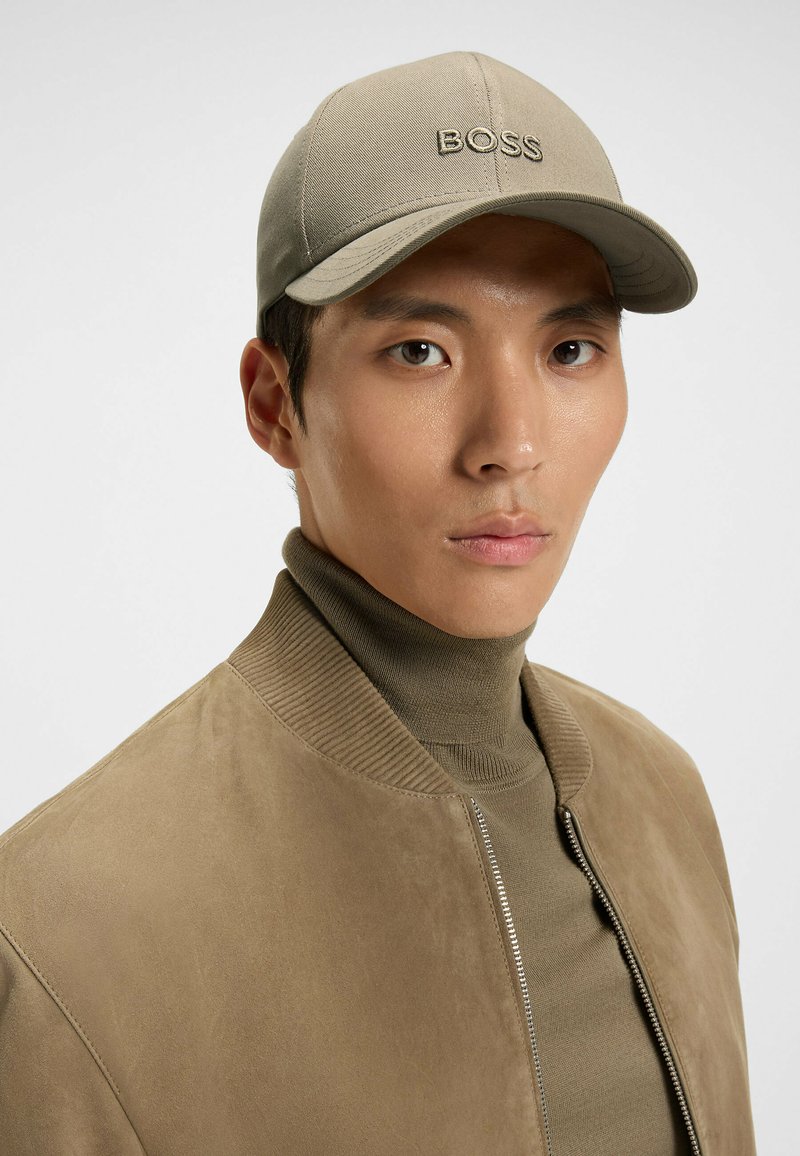 Khaki-baseballhatt med broderad "BOSS"-logotyp, bärs av en modell i en brun mocka bomberjacka över en beige polotröja.