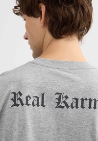 Camiseta gris de manga corta con gran texto gótico negro que dice "Real Karma" en la parte de atrás. Hecha de una tela suave y texturizada.