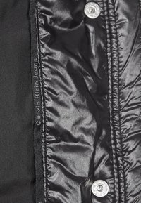 Svart puffjacka med glänsande inre, prydd med silverknappar och en svart etikett med "Calvin Klein Jeans" i kontrasterande sömnad.