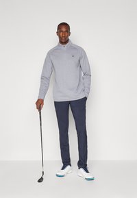 Grå zip-up golfpullover, mørkeblå bukser, hvite golf sko med blå detaljer, og en golfkølle. Glatt stoff med en avslappet passform.
