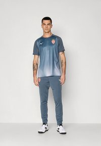 Sportieve outfit met een grijs T-shirt met verloopkleur en een teamlogo, gecombineerd met bijpassende joggers van gladde stof, en zwarte en witte schoenen.