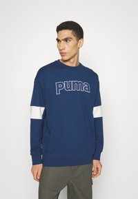 Navy blue sweatshirt made of cotton, featuring a large white "Puma" logo, white accent stripes on sleeves, and ribbed cuffs.