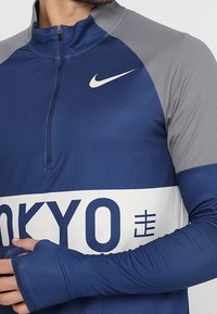 Man wearing a long-sleeve navy and gray Nike athletic shirt with "TOKYO" text and a quarter-zip collar, hands crossed at the waist.