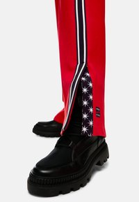 Red athletic pants with side zippers, featuring black and white stripes, a star-patterned lining, and paired with chunky black boots.