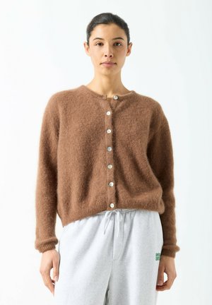 Kvinde iført en brun, blød, knappet cardigan og lysegrå joggingbukser med snor, stående mod en hvid baggrund.