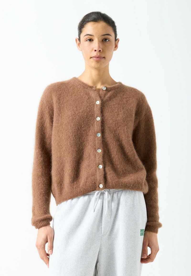 Sieviete, tērpusies brūnā, pūkainā pogājamā kardiganā un gaiši pelēkās sporta biksēs ar auklu, stāvot pret balto fona.