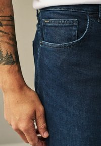 Tamsiai mėlynos džinsinės kelnės su artima nuotrauka šoninio kišenės ir rankos. Pavaizduota aiški siuvimo forma, subtilus blizgesys ir tatuiruota dilbis.