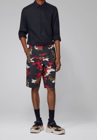 Muž v černé košili s knoflíky, červených, hnědých a bílých maskáčových šortkách, černých ponožkách a béžových s černými sandálech, stojící s rukama v kapsách.