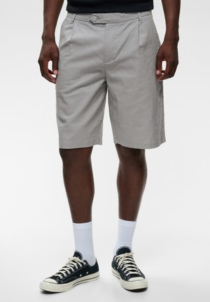 Man die lichtgrijze knielange shorts draagt, witte sokken en zwarte lage sneakers met witte veters, staande op een effen witte achtergrond.