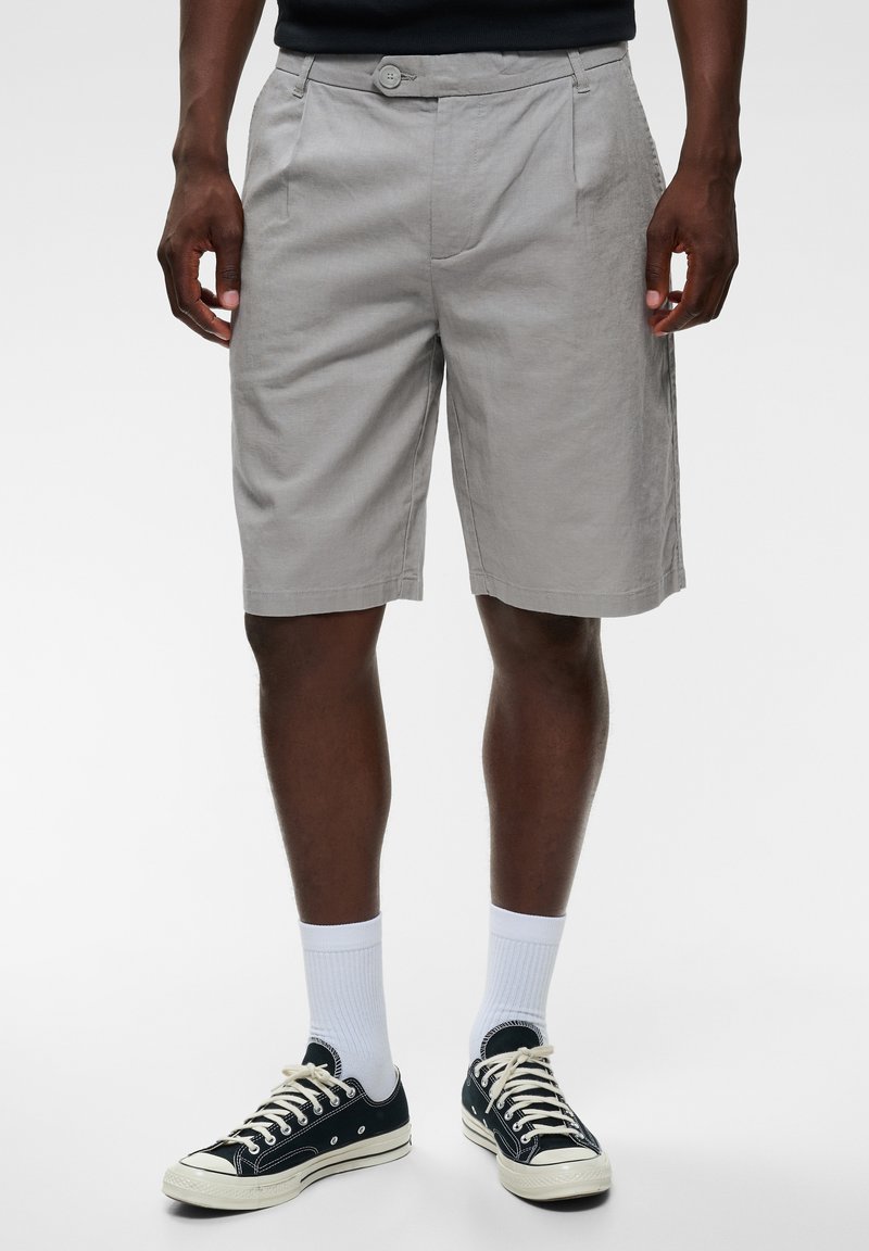 Homme portant un short gris clair jusqu'aux genoux, des chaussettes blanches et des baskets basses noires avec des lacets blancs, debout sur un fond blanc uni.