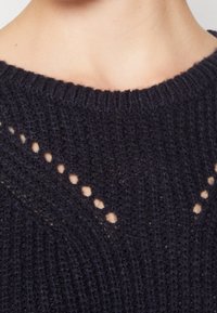 Marinblå stickad tröja med ribbat mönster och rund halsringning, med triangulära utskärningar längs framsidan och ett fint garnmönster.