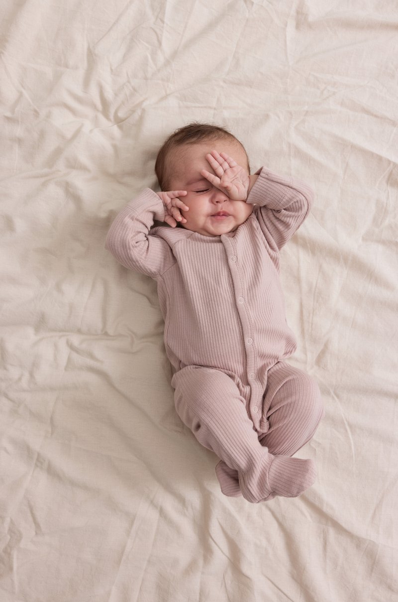 Lys rosa ribbestrikket heldrakt med lange ermer og knapper foran på et sterkt krøllet beige teppe; en baby leker med å dekke øynene med hendene.