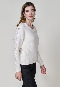 Weißer Strickpullover mit V-Ausschnitt, der Spitzenmuster entlang der Ärmel und gerippten Säumen aufweist. Kombiniert mit schwarzen, engen Hosen.