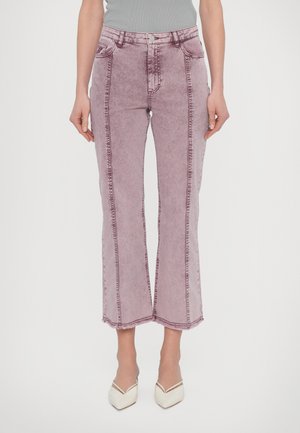 Kobieta ubrana w jasnofioletowe, wysokowalne jeansy typu flare z wykończeniem szwów i białe szpilki z zaostrzonym noskiem, stoi na tle jednolitym.