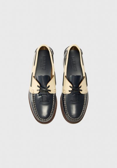 Scarpe da barca in pelle con una combinazione di colori blu navy e beige, punta tonda, design con lacci e cuciture a contrasto su una suola marrone.