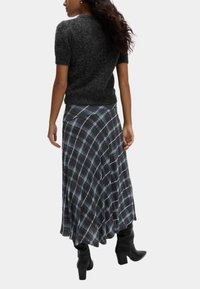 Dunkelgrauer, kurzärmeliger Strickpullover kombiniert mit einem langen, karierten Rock in Schwarz- und Blautönen, ergänzt durch schwarze Absatzstiefel.