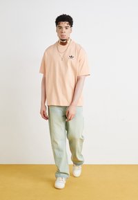 T-shirt oversized de pêssego claro com logótipo preto, acompanhado de jeans largos em verde menta desbotado e sapatilhas brancas. Design simples com mínimos detalhes.