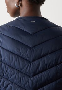 Personne portant une veste matelassée bleu marine avec un petit logo métallique sur le haut du dos, une boucle d’oreille argentée visible et des cheveux courts et bouclés.