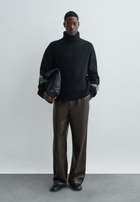 Črn pulover z visoko ovratnikom in zelenim cvetličnim vezenjem na rokavih, v kombinaciji z rjavimi hlačami in držanjem črne torbice. Gladke teksture.