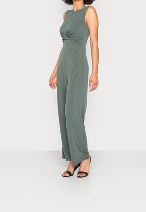 Femme portant une combinaison sans manches vert olive avec un détail torsadé à l'avant et des talons ouverts noirs, debout devant un fond uni.