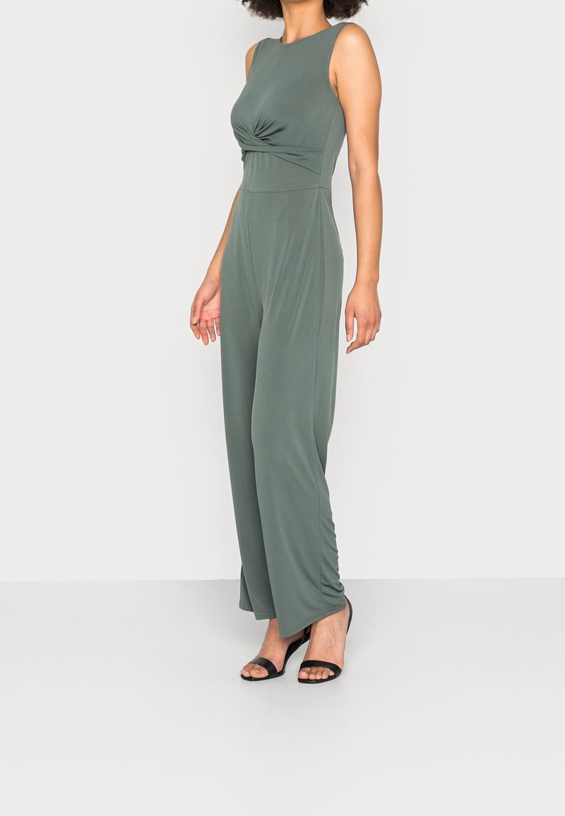 Femme portant une combinaison sans manches vert olive avec un détail torsadé à l'avant et des talons ouverts noirs, debout devant un fond uni.