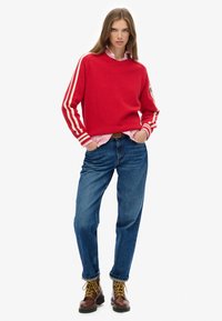 Vrouw in een rood sweatshirt met witte strepen, een roze gestreept shirt, blauwe jeans en bruine veterschoenen, staand met handen in de zakken.