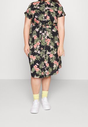 Persoon draagt een zwarte jurk tot op de knie met roze en groene bloemenprints, gecombineerd met gele sokken en witte sneakers, staand op een grijze vloer.