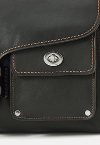Black leather bag with contrasting brown stitching, silver hardware, front pocket, and a flap closure. Smooth texture with a polished finish.
