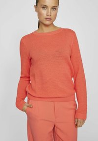 Korallfärgad stickad tröja med ett strukturerat mönster, rund halsringning och ribbade muddarn. Kombinerad med matchande korallbyxor för en enhetlig look.