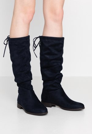 Vue en longueur de jambe d'une personne portant des bottes souples en suède bleu marine foncé montant jusqu'aux genoux, avec des petits talons et un détail de lien à l'arrière, sur un sol gris.