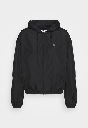 Chaqueta negra ligera con capucha, que cuenta con un cierre frontal, puños elásticos y un pequeño detalle del logo en el pecho. Textura suave y mate.