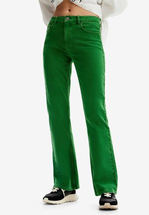 Jean flare en denim vert avec poches avant et passants de ceinture. Ourlet fini et tissu texturé. Associé à des baskets noires et crème.