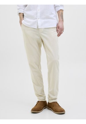 Mann iført off-white bukser, hvit skjorte med knapper og oppbrettede ermer, brune semskede sko og synlige tatoveringer på armene, stående med hendene delvis i lommene.