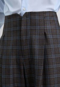 Los pantalones de cuadros marrones presentan líneas azules, un diseño de cintura alta y pliegues marcados. La tela tiene un acabado texturizado, lo que realza la profundidad del patrón.