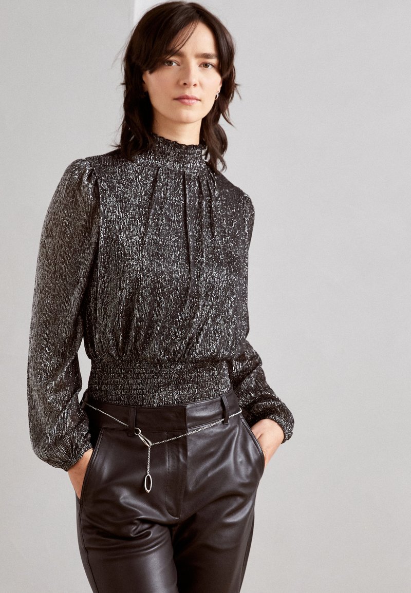 Blouse noire ornée de paillettes avec col haut, devant plissé et manches froncées. Associée à un pantalon en cuir noir avec un détail en chaîne.