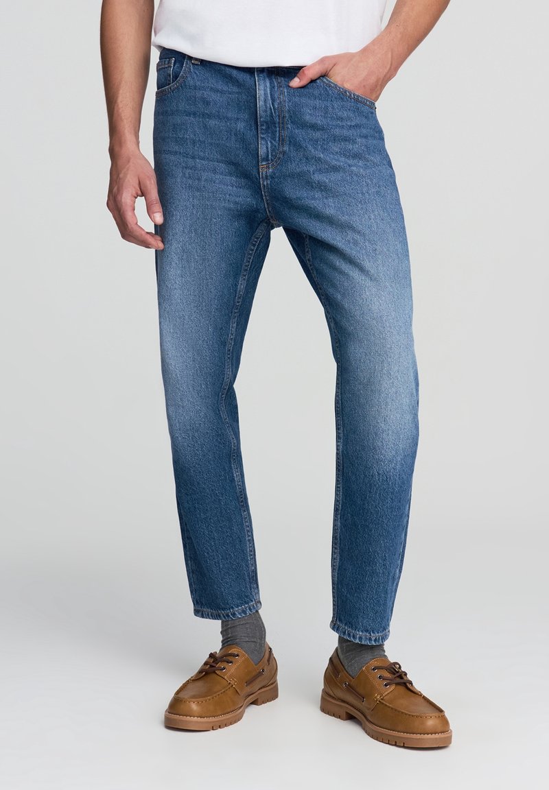 Hombre con jeans de mezclilla azul, calcetas grises y zapatos de cuero marrón con cordones, de pie sobre un fondo claro.