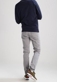 Felpa blu, jeans grigi slim-fit e sneaker olive con accenti arancioni. L'outfit casual sottolinea il comfort e il design moderno.