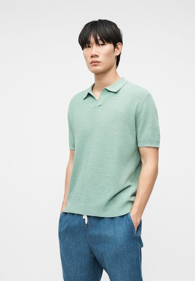 Νέος άνδρας με μαύρα μαλλιά, φορώντας ανοιχτοπράσινο πόλο με κοντά μανίκια και μπλε παντελόνι με κορδόνι, στέκεται με τα χέρια στις τσέπες.