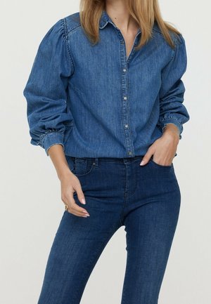 Frau trägt ein blaues Denim-Hemd mit Puffärmeln und dunklen, engen Jeans, eine Hand in der Tasche, vor einfachem Hintergrund.