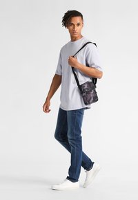 Czarna torba na ramię w kamuflażu, z przednią kieszenią na zamek, wykonana z trwałego materiału, o smukłym designie; noszona z jasnoszarym T-shirtem i niebieskimi dżinsami.