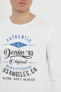 Bijela majica dugih rukava s okruglim izrezom, na kojoj su ispisani plavi i crni tekst, uključujući "Autentični Denim osnovan 93" i "Los Angeles, CA."