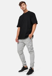 Mann trägt ein schwarzes, weit geschnittenes T-Shirt, hellgraue Jogginghosen und schwarze Sneakers, steht mit einer Hand in der Tasche vor weißem Hintergrund.