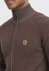 Brauner Reißverschluss-Pullover mit geripptem Kragen, strukturiertem Stoff und einem Lederpatch mit Logo. Akzente aus helleren Fasern erzeugen einen gepunkteten Effekt.
