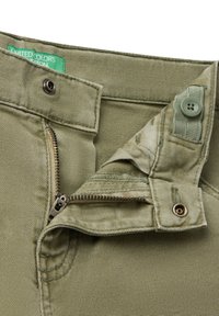 Dettaglio ravvicinato di pantaloni in tessuto verde aperti con bottone e chiusura a scatto, che mostra l'etichetta United Colors of Benetton all'interno della cintura.
