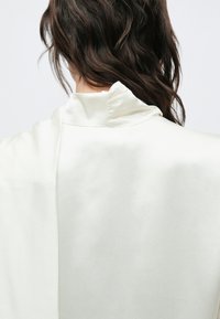 Jedwabna bluzka w delikatnym białym kolorze o minimalistycznym designie, z wysokim kołnierzem i gładką fakturą bez widocznych zapięć.