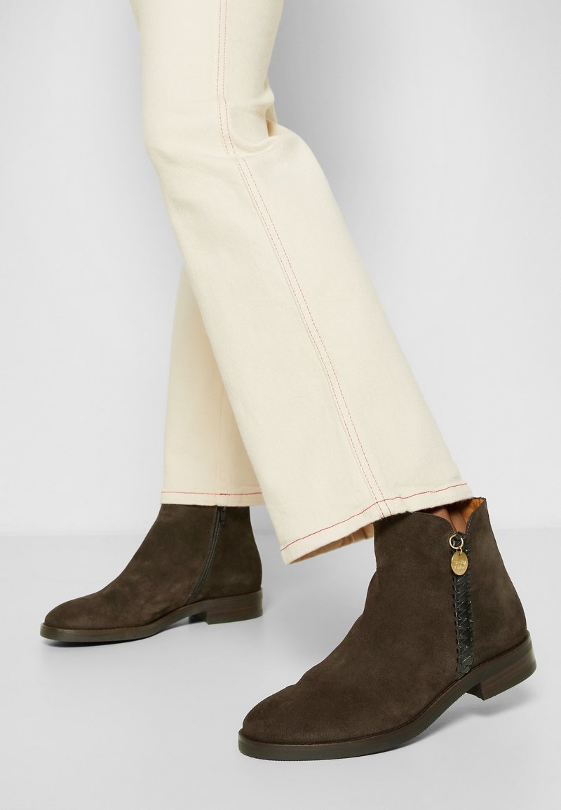 Bottes basses en daim marron avec fermeture éclair latérale et détail tressé décoratif, portées avec un pantalon crème à coutures apparentes.