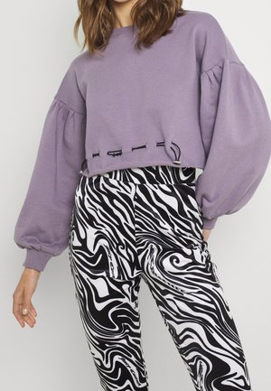 Person iført en lilla cropped sweatshirt med ballonærmer og sorte og hvide bukser med abstrakt mønster.