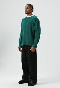 Smaragdgroene gebreide trui met lange mouwen, ronde halslijn en een losse pasvorm, gecombineerd met zwarte broek met brede pijpen en zwarte schoenen met veters.